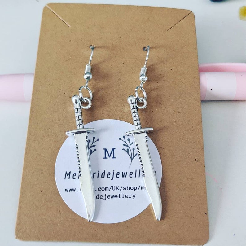 May include: Silver sword earrings on a brown card with the text "Merridejewellery.com/UK/shop/merridejewellery" and the letter "M" in the center.