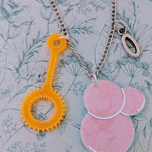 Puede incluir: Collar de cadena plateada con dos colgantes. Uno es una varita de burbujas amarilla, y el otro es un diseño de burbujas rosa con un estampado floral. El fondo es un diseño floral.