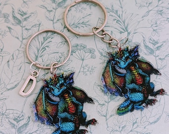 May include: Two dragon keychains with silver key rings. The dragons are blue with rainbow wings and accents. One keychain has a silver letter "D" charm. The keychains are on a floral patterned background.