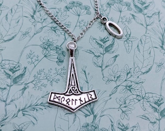 May include: A silver necklace with a Mjolnir pendant. The pendant has a knot design and the word "Mjolnir" inscribed on it. The necklace also has a small silver circle charm.