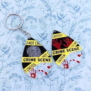 Può includere: Un paio di orecchini a forma di lacrima neri e bianchi con un design di nastro di scena del crimine giallo e nero. Gli orecchini presentano impronte di mani rosse e il testo "ATTENZIONE! SCENA DEL CRIMINE".