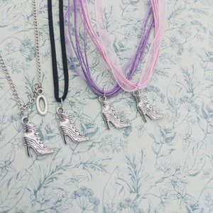 May include: Four silver boot charm necklaces with different colored ribbon chains. The chains are purple, pink, black, and silver. The charms are all the same design, a silver boot with a lace-up front.