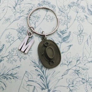 May include: A silver keychain with a letter M charm and a green hat charm. The keychain is on a floral patterned background.