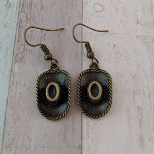 Könnte beinhalten: Ein Paar bronzefarbene Ohrringe mit einem Cowboyhut-Design. Die Hüte sind mit einer schwarzen und goldenen Oberfläche verziert.