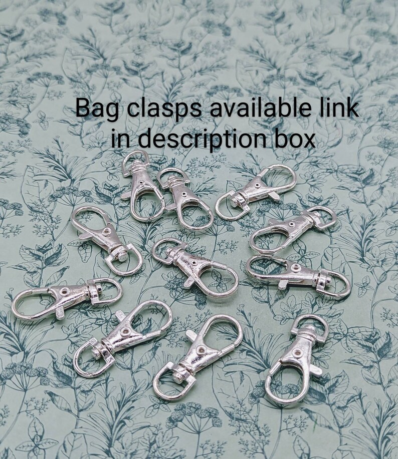 May include: A close-up of twelve silver-toned metal bag clasps, also known as lobster clasps, on a green floral patterned background.  The clasps are arranged in a random pattern.  Bag clasps available link in description box.