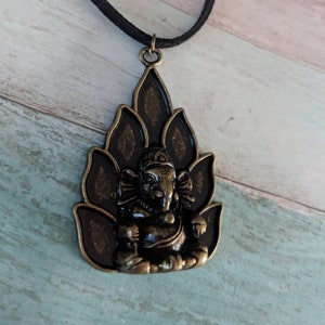 May include: A black cord necklace with a bronze pendant featuring a detailed depiction of the Hindu deity Ganesha sitting on a lotus flower.