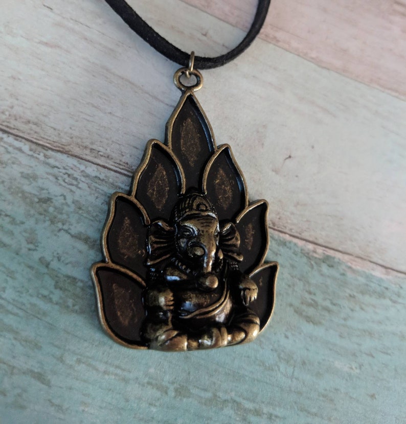 May include: A black cord necklace with a bronze pendant featuring a detailed depiction of the Hindu deity Ganesha sitting on a lotus flower.