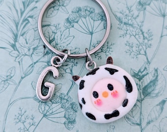 May include: A silver keyring with a cow-shaped charm and a letter "G" charm. The cow charm is white with black spots, brown ears, and a pink face. The keyring is on a light blue floral background.