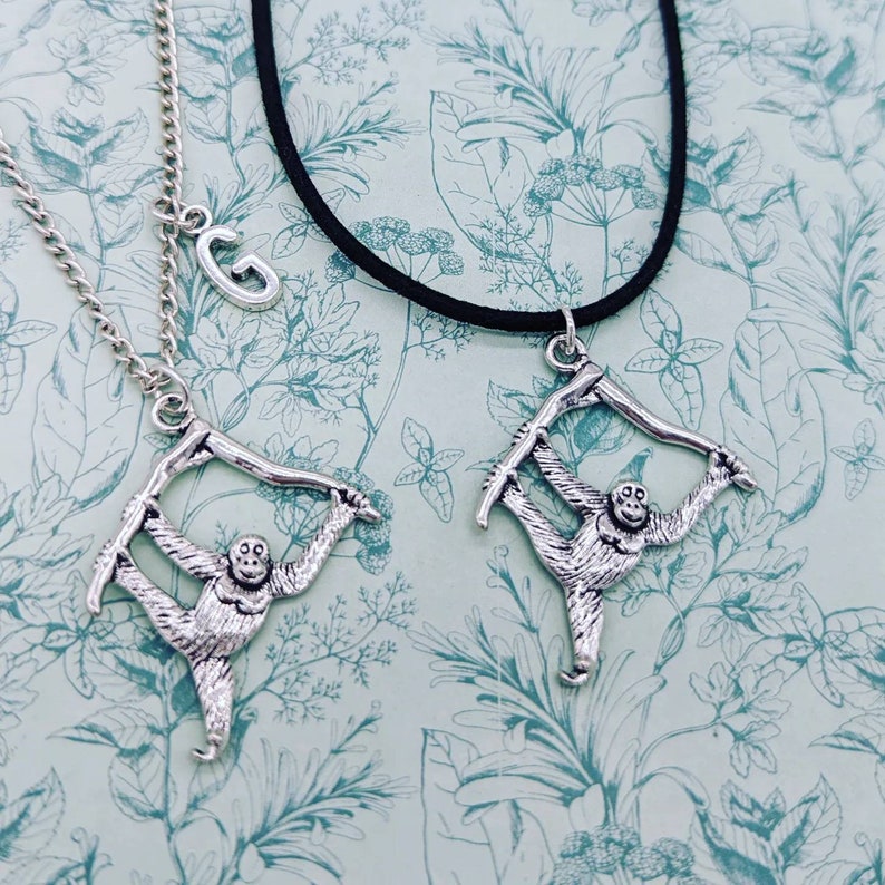 May include: Two silver necklaces with a monkey pendant hanging from each. One necklace has a silver letter G charm. The necklaces are on a floral patterned background.