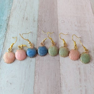 Op de afbeelding: Zes paar gouden oorhangers met een schelpdesign. De schelpen zijn in tinten blauw, groen en roze.