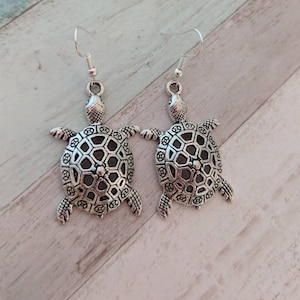 Puede incluir: Un par de pendientes de tortuga plateados con un diseño detallado e intrincado en la concha. Los pendientes cuelgan de ganchos plateados.