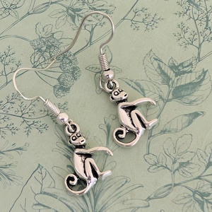 Op de afbeelding: Een paar zilverkleurige bungelende oorbellen met een kleine apenhanger. Elke aap is afgebeeld in een zittende positie met een gekrulde staart en gedetailleerde gelaatstrekken. De bedels zijn bevestigd aan standaard zilverkleurige oorhaakjes.