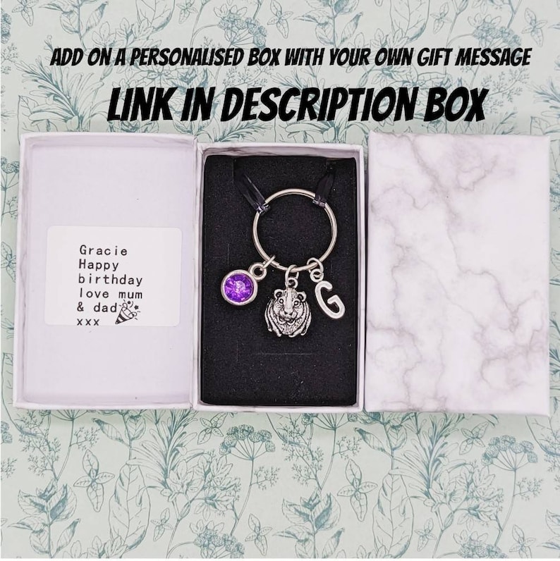 May include: A silver keychain with a guinea pig charm, a purple gem, and the letter "G" in an open gift box. The box contains a personalized message. Text on the image reads "ADD ON A PERSONALISED BOX WITH YOUR OWN GIFT MESSAGE LINK IN DESCRIPTION BOX."