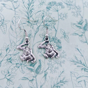 Puede incluir: Un par de pendientes de mono plateados. Cada pendiente presenta un dije de mono detallado, sentado, con el brazo levantado hacia la cabeza, suspendido de un gancho simple. Los pendientes se muestran sobre un fondo verde claro con un patrón floral.