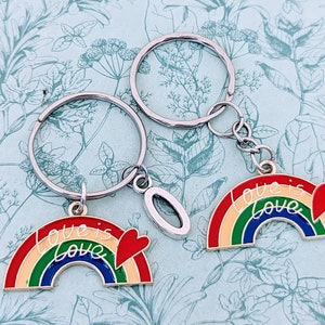 May include: Two silver keychains with rainbow-colored enamel charms. The charms are shaped like rainbows with the words "Love is Love" written on them. The keychains are a great gift for couples or friends.