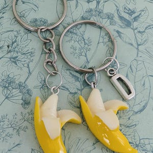 Op de afbeelding: Twee sleutelhangers met zilveren ringen en kettingen. Elke sleutelhanger heeft een gele bananenbedel met een witte binnenkant. Eén sleutelhanger heeft een rechthoekige zilveren bedel.