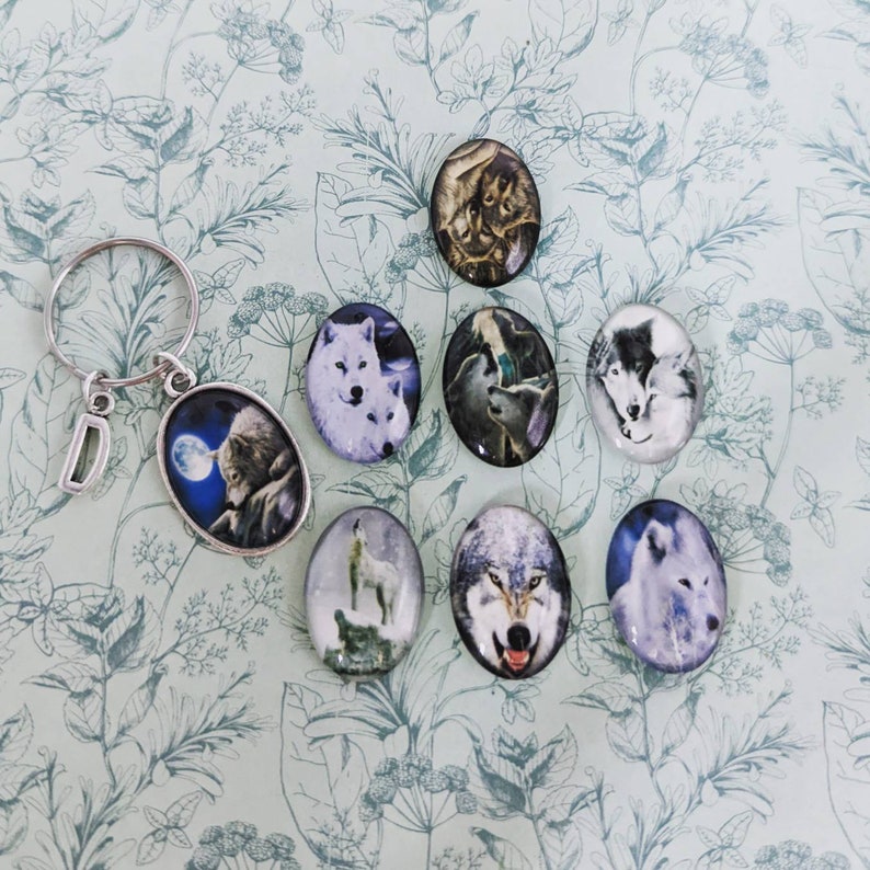 May include: A set of eight oval-shaped glass cabochons featuring images of wolves. The cabochons are silver-toned and have a clear glass finish. The images depict wolves in various poses, including howling, running, and cuddling.