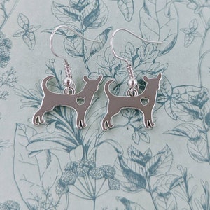 May include: A pair of silver dangle earrings featuring a silhouette of a chihuahua with a small heart cutout. The earrings are on a floral patterned background.