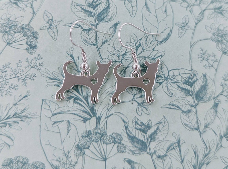 May include: A pair of silver dangle earrings featuring a silhouette of a chihuahua with a small heart cutout. The earrings are on a floral patterned background.