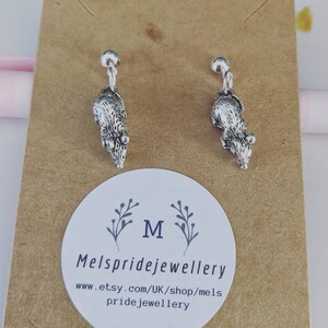 May include: A pair of silver rat earrings with a clip-on fastening. The earrings are displayed on a brown card with the text "Mels pridejewellery" and "www.etsy.com/UK/shop/mels pridejewellery".