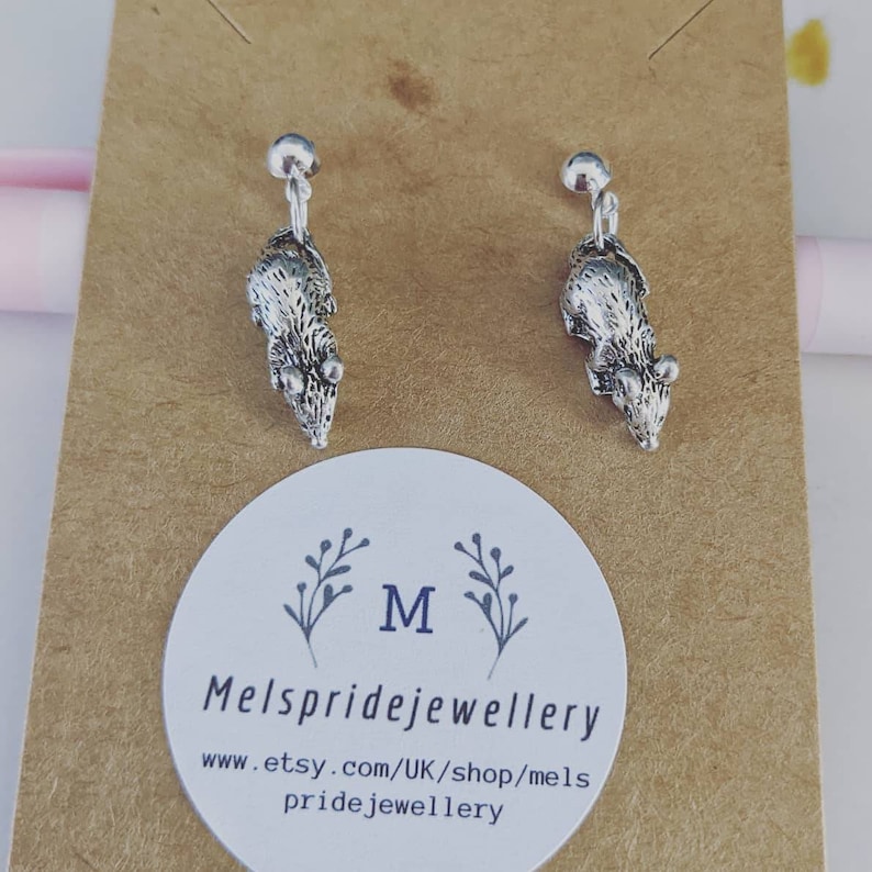 May include: A pair of silver rat earrings with a clip-on fastening. The earrings are displayed on a brown card with the text "Mels pridejewellery" and "www.etsy.com/UK/shop/mels pridejewellery".