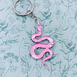 May include: Pink snake keyring with a silver key ring. The snake is decorated with white stars, moons, and other celestial designs.