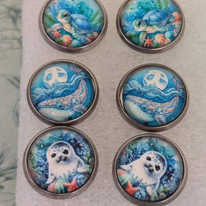 May include: Six round, silver-toned buttons with ocean-themed designs. Two buttons feature a sea turtle, two show a whale under a full moon, and two depict a seal. The artwork is in shades of blue, green, and coral.