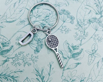 May include: A silver key chain with a tennis racket charm and a letter D charm. The charms are attached to a silver key ring.