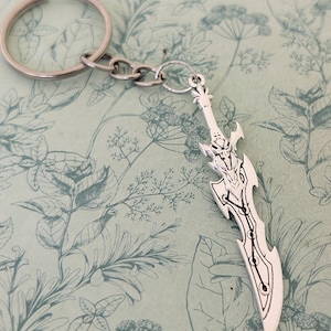 May include: Silver-colored sword-shaped keychain with intricate detailing. The keychain features a decorative sword pendant with a detailed hilt and blade design. The keychain is attached to a silver key ring and chain.