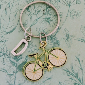 May include: A silver keyring featuring a green bicycle charm with white wheels and black detailing. A silver letter "D" charm is also attached. The background is a light green paper with a botanical design.
