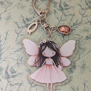 May include: A keyring featuring a clear acrylic charm of a fairy with dark hair and a pink dress, attached to a silver-toned metal ring. The keyring includes a small letter Q charm and a single, faceted, teardrop-shaped peach-coloured crystal charm.