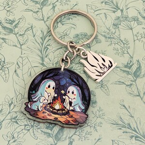 May include: A keychain with a silver ring and two charms. One charm is a clear, round acrylic with a cartoon scene of two ghosts and a dog around a campfire. The other charm is a silver, stylized flame.