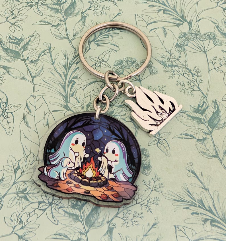May include: A keychain with a silver ring and two charms. One charm is a clear, round acrylic with a cartoon scene of two ghosts and a dog around a campfire. The other charm is a silver, stylized flame.