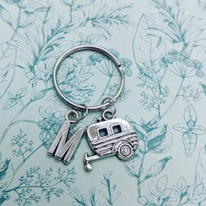 May include: Silver keychain with a letter M charm and a camper charm. The charms are attached to a silver key ring.
