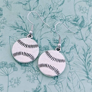 Peut inclure: Une paire de boucles d'oreilles en argent avec un motif de baseball. Les boucles d'oreilles sont sur un fond bleu clair avec un motif floral.