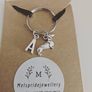 K&ouml;nnte beinhalten: Silberner Schl&uuml;sselanh&auml;nger mit einem Buchstaben A-Anh&auml;nger und einem Dackel-Anh&auml;nger. Der Schl&uuml;sselanh&auml;nger befindet sich auf einer braunen Karte mit dem Text "Mels pridejewellery" und einem kleinen Blumenmuster.