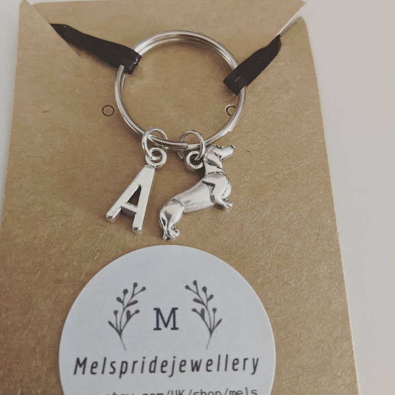 K&ouml;nnte beinhalten: Silberner Schl&uuml;sselanh&auml;nger mit einem Buchstaben A-Anh&auml;nger und einem Dackel-Anh&auml;nger. Der Schl&uuml;sselanh&auml;nger befindet sich auf einer braunen Karte mit dem Text "Mels pridejewellery" und einem kleinen Blumenmuster.