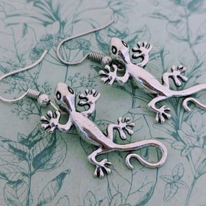 May include: A pair of silver dangle earrings featuring a detailed gecko design. The geckos are facing opposite directions and have textured bodies.