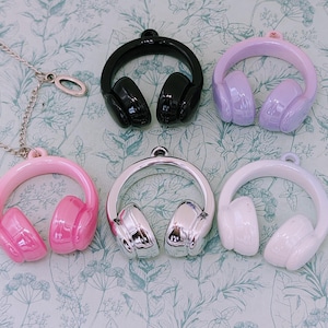 Könnte beinhalten: Eine Sammlung von Kopfhörer-Anhängern in verschiedenen Farben, darunter Rosa, Schwarz, Silber, Weiß und Lavendel. Ein rosa Kopfhörer-Anhänger ist an einer silbernen Halskette befestigt. Die glänzenden Anhänger haben eine Schlaufe zum Befestigen an Schmuck.