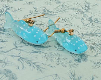 May include: A pair of earrings shaped like whale sharks. The earrings are light blue with white polka dots and gold-colored accents. The earrings are on a floral patterned background.