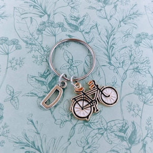 May include: Silver-tone keychain with a bicycle charm and a letter "D" charm. The bicycle charm is black and gold with white wheels. The keychain is on a light blue background with a floral pattern.