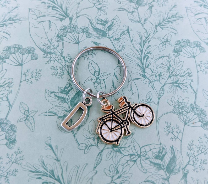 May include: Silver-tone keychain with a bicycle charm and a letter "D" charm. The bicycle charm is black and gold with white wheels. The keychain is on a light blue background with a floral pattern.