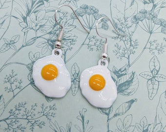 May include: A pair of novelty earrings featuring a fried egg design. Each earring has a white enamel egg white with a bright yellow yolk. The earrings hang from silver-colored fish hook ear wires.