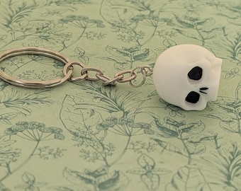 May include: A white skull keychain with black eye sockets and a black mouth. The keychain is attached to a silver chain and a silver ring.