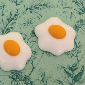 May include: Two handmade, star-shaped fried egg decorations. The eggs have white 'whites' and bright yellow-orange yolks. The wavy-edged eggs are set against a green background with a botanical pattern.