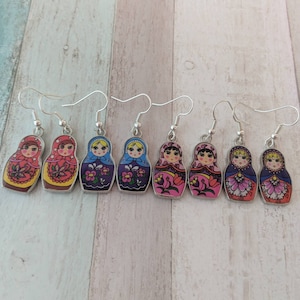 May include: A set of six pairs of earrings featuring a Russian nesting doll design. The dolls are painted in various colors, including red, blue, pink, and yellow. Each doll has a different facial expression and floral pattern.