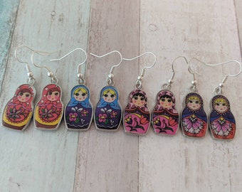 May include: A set of six pairs of earrings featuring a Russian nesting doll design. The dolls are painted in various colors, including red, blue, pink, and yellow. Each doll has a different facial expression and floral pattern.