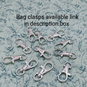 May include: A close-up of twelve silver-toned metal bag clasps, also known as lobster clasps, on a green floral patterned background.  The clasps are arranged in a random pattern.  Bag clasps available link in description box.