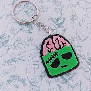 May include: A green and pink keychain featuring a cartoon brain with a stitched-up face. The keychain is attached to a silver key ring.
