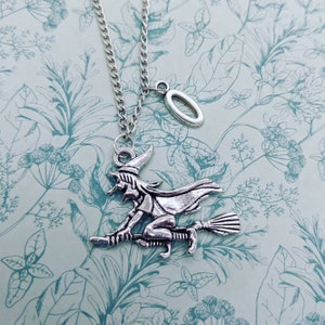 Puede incluir: Un collar de plata con un dije de una bruja montando una escoba y un dije ovalado de plata. El collar está sobre un fondo azul claro con un patrón floral.